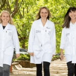 Four women scientists, in white coats, in front of green trees in a clinic, laughing and walking towards the camera.