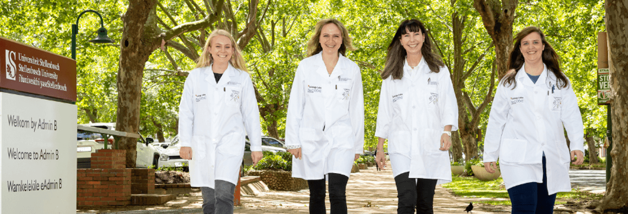 Vier Wissenschaftlerinnen, im weißen Kittel, vor grünen Bäumen einer Klinikanlage, lachend auf die Kamera zulaufend.