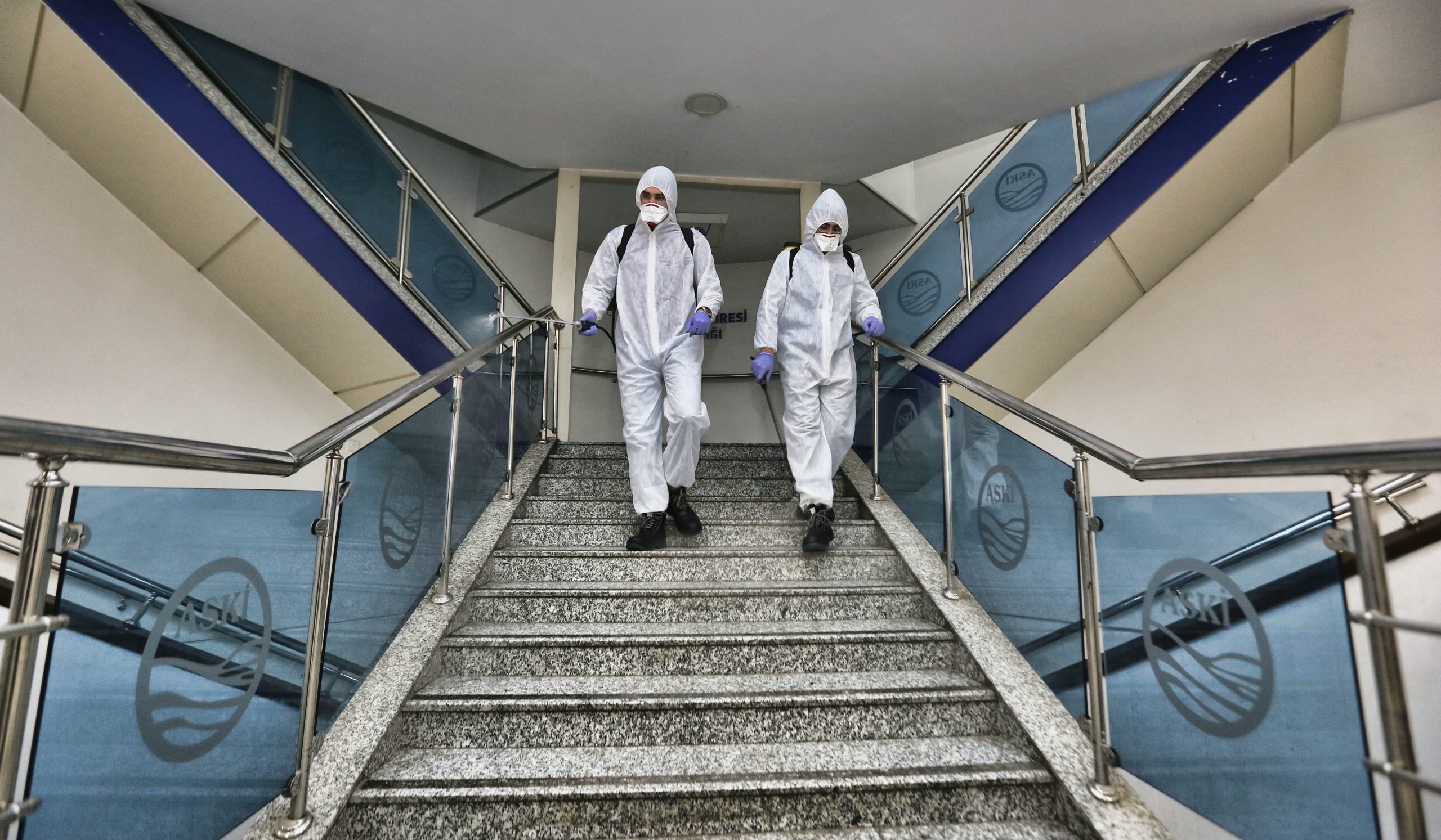Zwei Menschen in biologischer Schutzkleidung gehen eine Treppe hinunter.